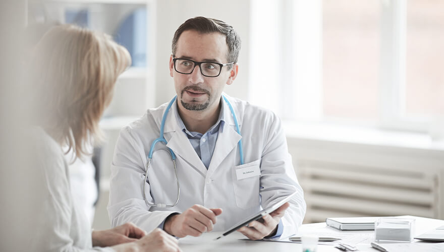 Arzt mit Brille und Stethoskop spricht mit Patientin am Tisch.