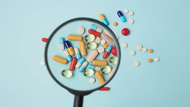 Verschiedene bunte Pillen und Tabletten liegen auf hellblauem Hintergrund, durch eine Lupe betrachtet.